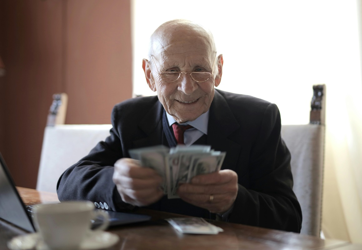 Usmívající se starší muž v obleku přepočítává svazek dolarových bankovek u stolu